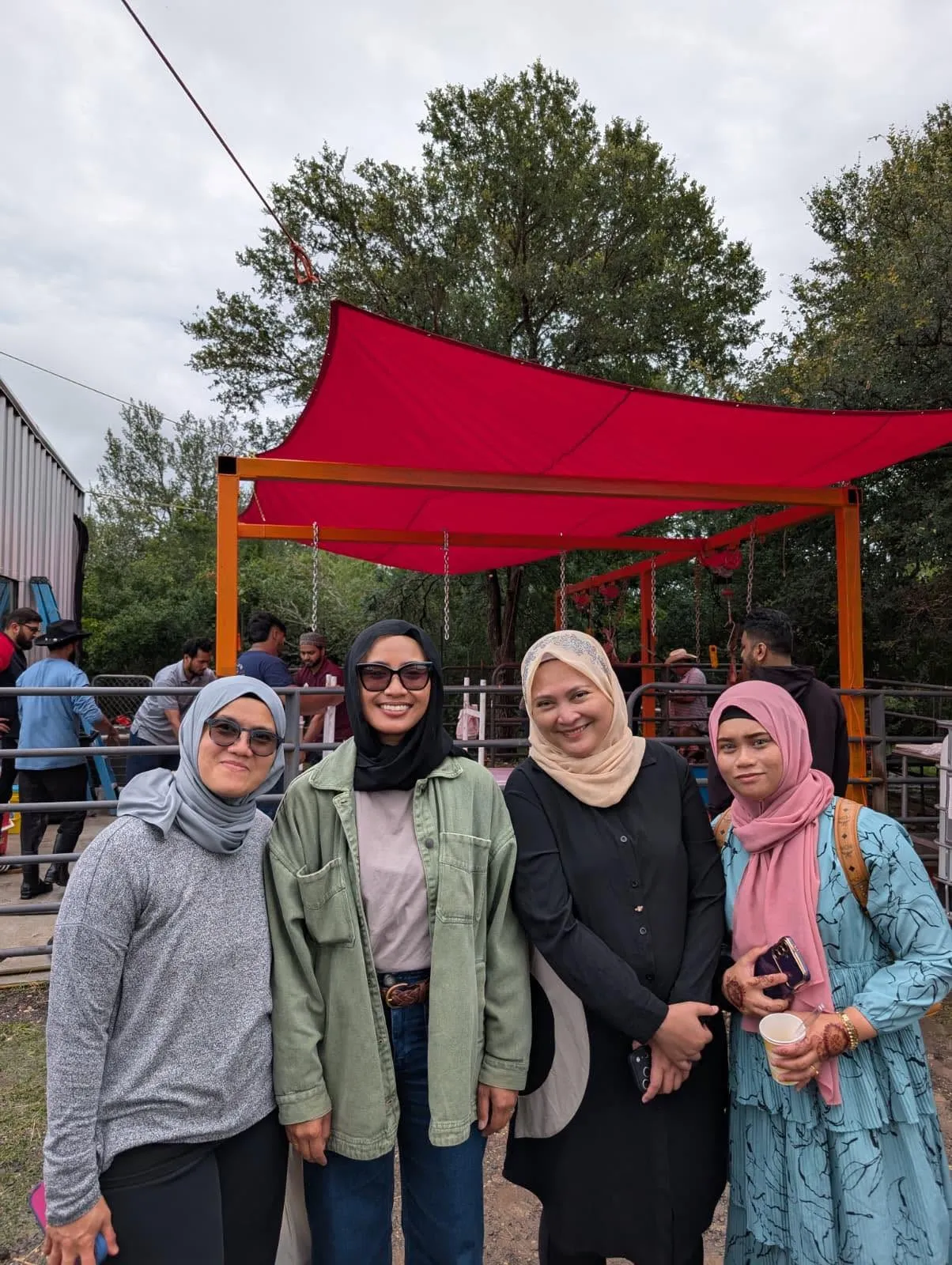 Four sisters smiling at the farm, with the red shade canopy of the qurban area behind them
