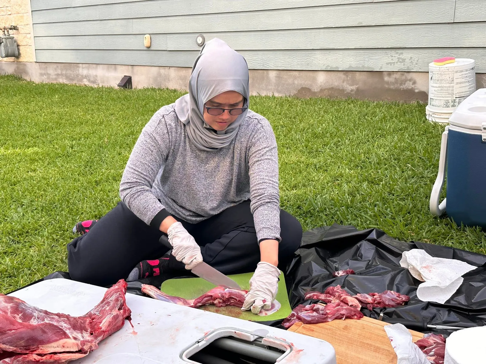 A sister in gloves cutting qurban meat into portions on a cutting board in a backyard